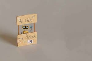 A small robot peeks out from behind a wooden stand with signs reading "AI Chat" and "The chatbot is IN".