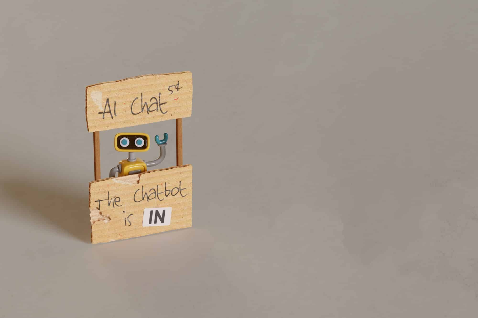 A small robot peeks out from behind a wooden stand with signs reading "AI Chat" and "The chatbot is IN".